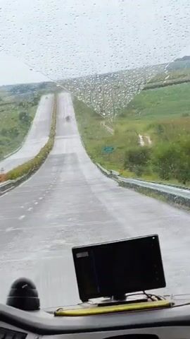 The video is filmed from the perspective of someone inside a vehicle, looking through the windshield. It is raining, and raindrops are visible on the glass. The road ahead is wet and appears to be a highway or a long, winding country road with a slight uphill incline. Greenery, possibly fields or hills, lines the sides of the road. There are guardrails along the sides of the road. In the foreground, on the dashboard of the vehicle, is a black electronic device, possibly a GPS or a display screen, with a green indicator light. Further down the road, a few other vehicles are visible in the distance. The sky appears overcast.