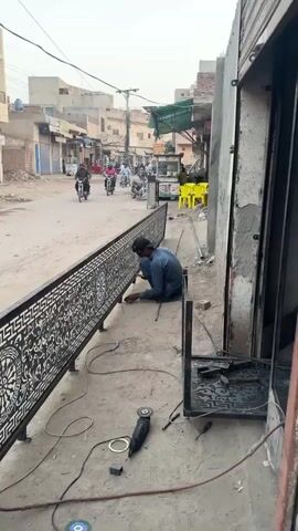 In Nasheman Colony, Pakistan, a man wearing a blue shalwar kameez and a black cap is crouched on the ground, actively engaged in working on a long, black metal fence or railing. The fence features intricate white laser-cut patterns, including floral motifs and a Greek key border, and appears to be in the process of installation or repair alongside a building. Welding equipment, including a torch or welding rods and an angle grinder, are visible on the dusty ground near the worker.

In the background, the scene depicts a typical street in a developing urban or semi-urban area, with several people riding motorcycles along the unpaved road. Buildings, possibly houses or shops, line both sides of the street, and power lines crisscross overhead. Yellow plastic chairs are visible on the right side of the street, suggesting an outdoor seating area. The weather appears clear, and it is daytime, likely late morning or afternoon. The man is focused on his task, and the overall atmosphere is one of routine daily activity in the neighborhood.