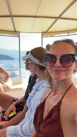 The video shows a group of people on a boat. In the foreground, a woman with sunglasses and her hair in two buns smiles at the camera. Behind her, a person wearing a white visor and sunglasses looks away from the camera, and another person sits partially visible on the left. The boat appears to be moving on the water, with islands and hills visible in the background. The sun is shining, casting shadows and illuminating the scene.