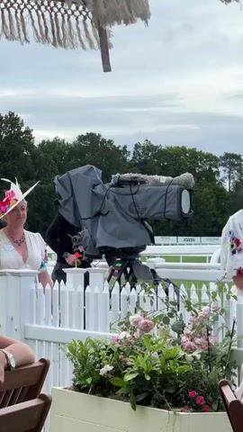 A woman in a white dress with colorful floral embroidery and a wide-brimmed white hat decorated with pink and orange flowers stands near a large camera. The camera, covered in a grey protective casing, is mounted on a tripod. To the right of the camera, another woman in a navy blue dress and a black hat speaks to someone off-camera. In the background, a white picket fence separates the foreground from a green lawn and trees. A planter filled with pink roses and other greenery sits in front of the fence. Another person in a green patterned outfit is visible behind the camera. The sky is overcast.