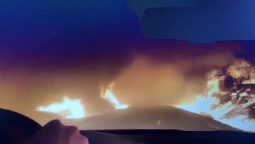 This video shows a person's hand on a steering wheel of a car. The view is from inside the car, looking through the windshield. Outside the car, there is a large fire with flames and smoke visible. The fire appears to be burning trees and brush. The sky is dark, and it looks like it is nighttime or dusk. The fire is very bright and casts a strong orange glow on the surroundings. The car appears to be driving through or near the fire.