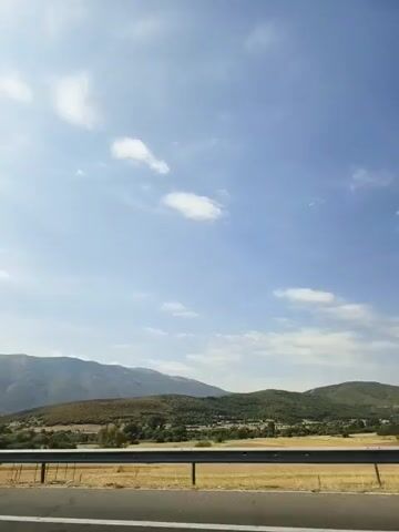 The video shows a car's perspective as it drives down a highway. The camera faces right, capturing the landscape on the side of the road. Mountains are visible in the distance, with some trees and fields closer to the highway. The sky is blue with scattered white clouds. There are no people or other vehicles visible in the video.