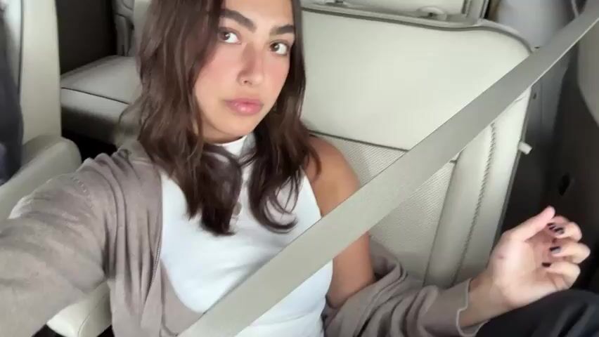 A woman with shoulder-length dark hair, wearing a white tank top and a beige cardigan, is sitting in the back seat of a car with beige leather seats. She is wearing her seatbelt and looks directly at the camera, giving the impression she is posing for a photo or video. The camera zooms in on her face before zooming back out to show more of the interior of the car. Another person is barely visible in the background, sitting next to her. Music is playing in the background.