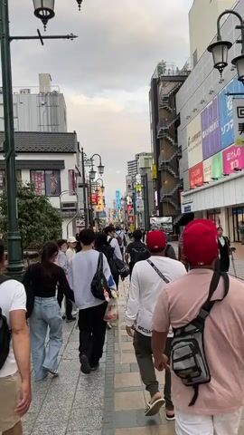 The video shows a street scene in an urban environment, likely in Asia given the architecture and signage. Several people are walking down a pedestrian pathway. In the foreground, there are four individuals walking away from the camera. The person furthest left wears a backpack and khaki shorts. Next to them, a woman in jeans and a dark top walks alongside a man in a white shirt and dark pants. Another person in a white shirt and red baseball cap walks slightly ahead and to the right. The street is lined with buildings, many of which have signs with Japanese text. Some buildings have multiple stories and appear to be commercial establishments. There are streetlights and electrical wires visible. The sky is overcast. The pathway is paved with interlocking bricks. The overall impression is that of a busy shopping district.