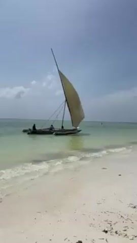 The video shows a beach scene with a traditional sailing boat anchored in shallow water. The boat has a large, triangular sail and appears to be a catamaran style with two narrow hulls. There are three people on board. The beach has white sand with some seaweed or debris scattered along the shoreline. The water is a light turquoise color, and small waves are lapping at the beach. The sky is bright with a few scattered clouds.
