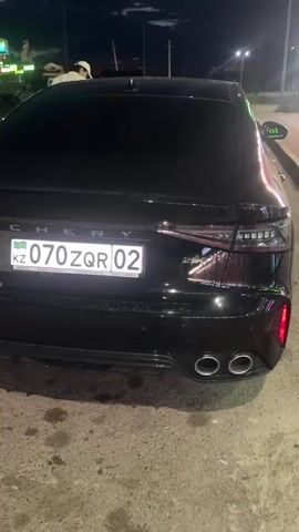 The video shows the rear of a black Chery Arrizo 8 car at night. The car has a license plate that reads "070 ZQR 02" with "KZ" visible on the left side. The car has four exhaust pipes. In the background, there are streetlights and a cloudy sky. A person is visible near the car in the background. The ground appears to be asphalt with some oil stains.