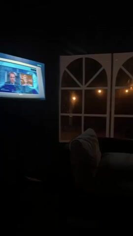 A television screen displays a news broadcast featuring two people, a man and a woman, against a blue background. The television is mounted on a dark wall. To the right of the television, a large window with arched panes looks out onto a dark yard. Lights are visible outside the window, illuminating a house with a pitched roof and multiple windows. A dark couch is partially visible in the foreground on the right. The overall scene is dimly lit.