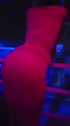 A woman in a tight pink dress leans against a railing in a dimly lit room, possibly a nightclub or bar, judging by the blue lights and music. The camera focuses on her back and posterior. The environment is dark, and the focus is on the woman and her dress.