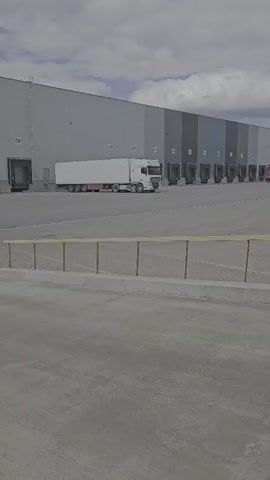 The video pans across a large industrial warehouse complex. Several trucks are parked in front of loading docks along the side of the building. The sky is cloudy.