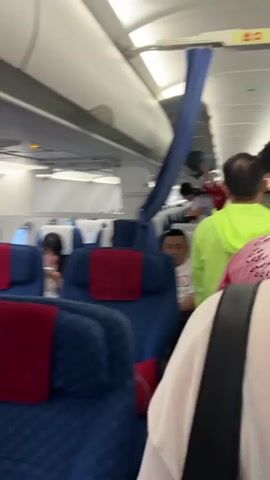The video shows the interior of an airplane aisle with passengers disembarking or boarding. Several people are visible standing and walking in the aisle, with luggage present. The seats are upholstered in blue with red headrests. An "EXIT" sign with Chinese characters above it is visible at the top of the frame. The lighting suggests it is daytime.