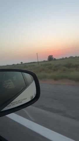 A car drives along a road. The view is from inside the car looking out the side window and in the side mirror. The side mirror reflects the driver's hand holding a cigarette and the interior of the car. Outside the car, a rural landscape of fields and a sunset are visible. The sky is a pale orange and pink. The sun is a small, distant orange orb. A single tree is visible in the distance. The road is paved and has white lines marking the lanes.