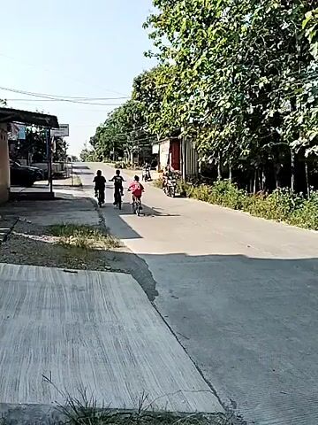 The video shows a street scene on a sunny day. A group of three individuals on bicycles are seen cycling away from the camera, followed by a black pickup truck with white sacks in its bed. The truck turns right. Later, a white scooter with a man wearing a dark t-shirt and light shorts rides past the camera in the opposite direction. The street is paved, and there are buildings and trees on both sides.