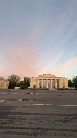 A vertical video begins with a wide shot of a classical building with columns and a long staircase leading up to it. The sky is a soft pink and blue with scattered clouds, indicating it is either dawn or dusk. The camera then pans to the right, showing a blurred view of other buildings and trees. It continues to pan and tilt upwards, the sky filling more of the frame. The video ends with a view of a monument with two statues in front of a tree-lined area, with a street lamp visible on the right. The sky is still brightly colored with pink and orange hues. The ground in the foreground appears to be wet, possibly from recent rain, with puddles visible.