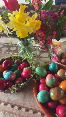 The video shows a table centerpiece with Easter decorations. There are two bowls filled with brightly colored Easter eggs in shades of red, green, blue, orange and brown. The eggs are speckled and arranged with sprigs of greenery. Two glass jars serve as vases, holding spring flowers including yellow daffodils, red tulips, and smaller pink and purple blossoms. The table is covered with a striped beige and brown cloth. A partially visible dog is on a couch in the background.