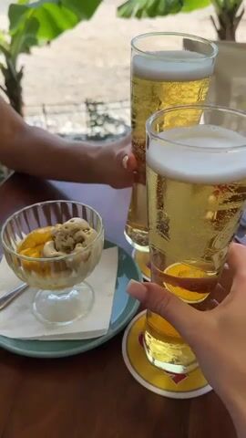 The video shows two hands holding tall glasses of beer, clinking them together in a toast. In the foreground, a glass bowl of ice cream with a yellow topping is visible. In the background, banana trees and a glimpse of a car and buildings can be seen, suggesting an outdoor dining setting on a sunny day. The overall atmosphere is relaxed and celebratory.