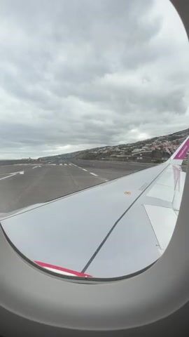 A view from inside an airplane window shows the plane approaching a runway for landing. The wing of the plane is visible in the foreground, and the runway stretches out in front. The runway is gray with white lines, and a building with red lights is visible in the distance. Beyond the runway, a hillside covered in houses and trees is visible. The sky is overcast and gray.