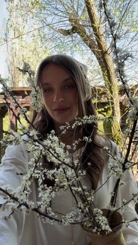 A young woman stands on a wooden bridge holding a vase with flowering branches. She's wearing a white shirt and a crochet headscarf. Behind her, there's a wooden pergola and trees against a blue sky. She looks at the camera and then glances to the side. There is some background noise, including snippets of speech.