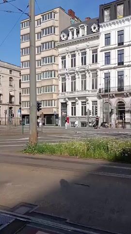 The video captures a street scene, likely in a European city, with a focus on tram tracks and overhead wires. In the foreground, tram tracks run across a paved surface, with some grass and greenery growing between them. A shadow of a person or object is cast on the ground. In the background, several buildings are visible, varying in architectural style. One building is a tall, modern structure with multiple floors and windows. Adjacent to it is a more ornate, older building with white facades and decorative elements. Further down the street, another older building with a rounded tower and mansard roof is visible. There are traffic lights, streetlights, and poles with overhead wires for the trams. Several people are seen walking or cycling, and a black car is driving on the road. The sky is clear and blue, indicating a sunny day. The overall impression is of an active urban environment with functioning public transportation.