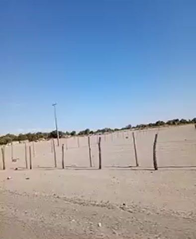 The video shows a bright, sunny, arid landscape with a clear blue sky. A sandy area is fenced with wooden posts, some of which are broken or leaning. Beyond the fence, sparse vegetation and scattered structures are visible. The camera appears to be moving, capturing the scene in motion. The scene appears desolate and sparse.