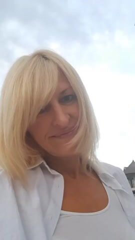 A woman with blonde hair and bangs smiles at the camera. She is wearing a white button-down shirt over a white tank top. The background is slightly blurred and appears to be outdoors with a cloudy sky and some buildings. The video is a selfie-style shot, with the camera positioned slightly above eye level.