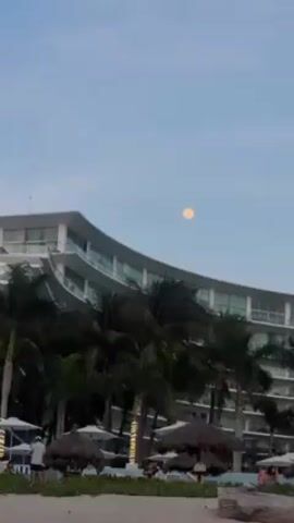 The video shows a beach scene, focusing on a large, yellowish moon in the sky. The camera pans upwards to emphasize the moon, which is the central subject. In the foreground, there's a white curved building, likely a hotel, partially obscured by palm trees. Below the trees, there are thatched-roof beach umbrellas and a few people, some standing and some relaxing in chairs. The beach appears sandy. The overall setting suggests a tropical resort atmosphere.