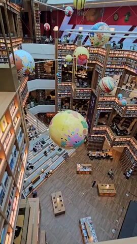 A high-angle shot pans across a multi-story bookstore with an atrium. Large, colorful spherical decorations, resembling planets or globes, are suspended from the ceiling. Bookshelves line the walls of each level, creating a maze-like structure. On various levels, people are seen sitting at tables, reading, and interacting. Stairs and walkways connect the different floors. In the center of the atrium, a large yellow globe with colorful splotches is prominently displayed. Wooden tables with seating arrangements are visible on the ground floor, suggesting a cafe or seating area. The overall ambiance is one of a spacious, modern, and inviting library or bookstore environment.