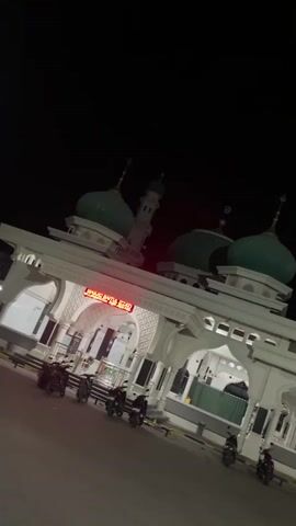 The video shows the entrance to a mosque at night. The mosque has several green domes and a minaret. The entrance is an archway with white ornate carvings. A red neon sign above the entrance reads "MASJID BAITUL IJAZAH". Several motorcycles are parked in front of the entrance. The sky is dark, indicating it is nighttime.