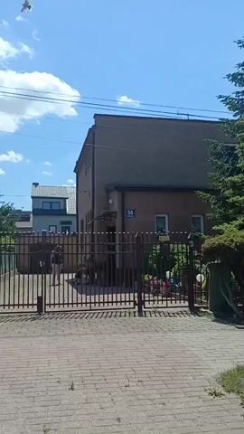 This media file shows a view of a building with a fence in front of it. The building is two stories tall and has a dark brown exterior. A blue sign with the number "54" is visible on the side of the building. There is a gate in the fence, and a person can be seen standing near the gate. The area in front of the fence is paved with bricks. Trees and plants are visible behind the fence. The sky is blue with some white clouds, suggesting it is daytime and the weather is clear. The overall setting appears to be a residential or commercial area in Ciechanów, Poland. There is also a yellow sign with text on it attached to the fence.