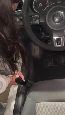 A young woman is seen inside of a car, operating a handheld vacuum to clean the floor. The driver’s side door is open. The car’s floor mats are dark gray, and the car seat is gray with a textured pattern. The steering wheel is black. The woman has long, dark hair, and she is wearing a light gray top.