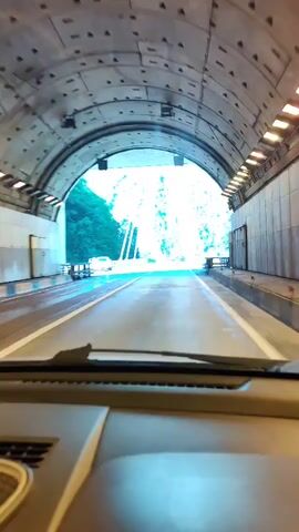 The video captures a car's point-of-view journey through a tunnel and onto a cable-stayed bridge. The initial shot shows the view from inside the tunnel, with the bright opening of the bridge visible ahead. The tunnel appears to be made of concrete, with visible construction elements. As the car exits the tunnel, the scene opens up to a bright, sunny day.

The prominent subject is a modern cable-stayed bridge, characterized by its tall, A-frame pylons and numerous cables supporting the roadway. The bridge spans a gorge or valley, with lush green, tree-covered mountains rising on either side. The roadway is asphalt with clear lane markings, and a guardrail runs along the sides. A white car is visible on the bridge in the distance. The sky is a clear, vibrant blue, suggesting fair weather.

The activity taking place is driving across the bridge. The time of day appears to be daytime, likely late morning or early afternoon, given the strong sunlight and bright conditions. The setting is mountainous and scenic, indicative of a natural landscape with significant infrastructure. The overall mood is one of progress and emergence into a bright, open space after the enclosed tunnel. There is no visible text in the video. The location is identified as Kepsha, Russia.