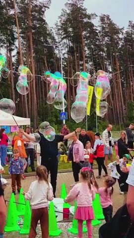 A wide shot shows a group of people, mostly adults and children, gathered around a street performer making bubbles. The performer, a man in a black suit with a bow tie and a green hat, is holding two sticks with strings between them to create large, colorful bubbles that float into the sky. The crowd watches with interest, and some children are reaching out to try and catch the bubbles. In the background, tall pine trees line the edge of the paved area, and a white tent is visible on the left. The sky is overcast.