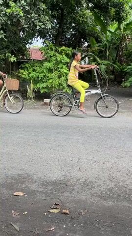 The video captures two young girls riding bicycles down a paved road lined with trees and modest buildings. In the initial frames, the girls are riding in the same direction, one slightly ahead of the other. A motorbike with two large, patterned cargo boxes attached to its rear passes them. The video then shifts to a wider view of the street, showing the girls continuing to ride, with the motorbike in the distance. The environment appears to be a village or rural area, with lush greenery and typical houses. The weather seems overcast. The girls are dressed casually, and the bicycles are simple models.