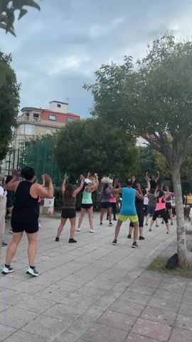 A group of people are doing a dance fitness class outdoors in a park on a paved walkway lined with trees. The group is facing away from the camera. One person in the front left is wearing a black tank top and shorts. Many others are wearing athletic wear in various colors. A building with red trim is visible in the background behind the trees. The sky is overcast.