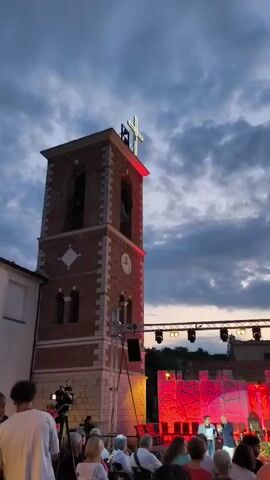 A short video shows a brick bell tower with a cross illuminated by white lights on top against a cloudy sky. Red lights are visible at the base of the cross and below the bell tower opening. A clock is visible on the tower. Below the tower, a crowd of people is gathered, some seated and some standing, watching a stage set up with red lighting and some structures that appear to be part of a performance. A camera operator is also visible in the foreground.