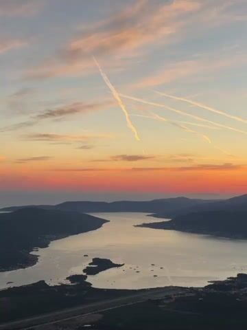 The video shows a wide shot of a bay at sunset. The sky is filled with streaks of orange, pink, and yellow clouds, with contrails from airplanes crisscrossing the upper atmosphere. Dark, silhouetted mountains surround the bay, which is filled with calm water reflecting the colors of the sky. The shoreline is visible on the left and right, with some lights suggesting a town or settlement. In the foreground, a dark railing is partially visible, indicating the viewer is on a balcony or observation deck. The overall impression is one of a serene and picturesque natural landscape during twilight.