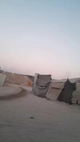 The video shows a ground-level perspective of a dusty, undeveloped area. The sky is a gradient of light blue and pink, indicating either dawn or dusk. In the foreground, there is a dirt road. To the right of the road, several makeshift structures are visible, constructed from what appears to be tarps or fabric draped over frames. These structures look like temporary shelters or stalls. Behind these structures, to the left, there is a pile of debris or excavated earth, with some sparse vegetation growing around it. There are no people clearly visible in the frame, and the overall impression is one of a rural or semi-arid environment with basic infrastructure.