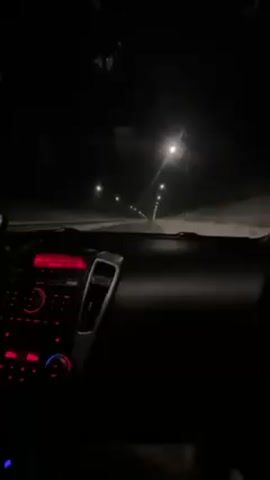 The video shows a car driving on a road at night. The road appears to be covered in snow. Streetlights illuminate the road ahead. The car's dashboard is visible, displaying red lights. The windshield has some water droplets or melted snow on it. The music is playing in the background. The overall environment is dark and cold.