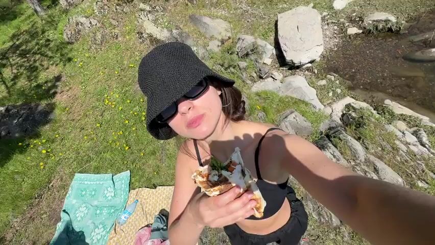 A young woman is prominently featured in a high-angle selfie shot, actively eating a large grilled wrap or kebab. She is wearing a black woven bucket hat, dark sunglasses, and a black strappy top. The wrap appears to be filled with ingredients like lettuce, meat, and sauce, with visible charred marks indicating it may have been recently grilled or toasted. She takes a large, enthusiastic bite, with some food remnants visible on her lip and chin afterward, suggesting she is enjoying her meal.

The setting is a natural outdoor environment under bright sunlight, indicating clear weather during the daytime. The ground beneath her is rocky and uneven, interspersed with patches of green grass and small yellow flowers. To her left, a dense forest of pine trees and other green foliage is visible. To her right, and slightly in the background, a calm body of water, likely a lake or river, can be seen with a light-colored object (possibly a log or debris) floating near the shore. The high camera angle suggests she might be on a cliff or elevated viewpoint overlooking the water, enjoying a meal amidst scenic nature.