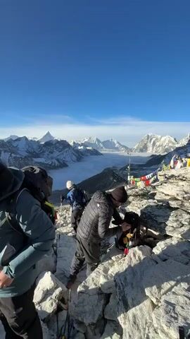 The video captures a group of trekkers in a stunning high-altitude mountain landscape under a clear blue sky.

**Subjects:** There are several people, appearing to be hikers or mountaineers, dressed in warm winter clothing including puffer jackets, beanies, and trekking pants. They carry backpacks and some have trekking poles.

**Objects:** Backpacks, phones (used for taking photos/videos), and colorful prayer flags tied to a pole are visible.

**Scene/Setting:** The setting is a rocky, uneven mountain ridge or viewpoint. In the background, a breathtaking panorama of snow-capped Himalayan peaks stretches across the horizon. A deep valley below is filled with a thick layer of white clouds or fog, creating a "sea of clouds" effect. The sun is bright, indicating a clear day, and casts some glare in certain frames. The specific location cues, including the distinctive mountain shapes and the general environment, strongly suggest the Everest region in Khumjung, Nepal. The peak of Mount Everest and Nuptse are prominently visible.

**Activity or Event:** The group appears to be resting, adjusting their gear, and actively taking photographs and videos of the spectacular scenery. One person in a yellow jacket strikes a pose while holding a phone out, seemingly for a photo or video. Another individual in a dark jacket is also holding a phone, possibly capturing the moment.

**Notable Details:**
*   **Time of day:** Bright daylight, with intense sun suggesting a morning or early afternoon.
*   **Weather:** Clear skies, sunny, and likely cold given the attire.
*   **Location cues:** The iconic snow-capped peaks of the Himalayas, including what appears to be Mount Everest and Nuptse, the rocky terrain, and the prayer flags, all point to a high-altitude viewpoint in the Everest region.
*   **Emotions:** The individuals seem to be in awe of the surroundings and enjoying the moment, as evidenced by their active engagement with photography.

**Visible Text:** There is no visible text in the video.