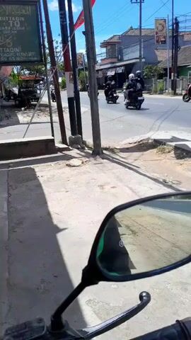 The video shows a street scene from the perspective of someone on a motorcycle. The camera is positioned to show the right side mirror, which reflects a green object and the road. In the background, there is a street with buildings on either side. Several motorcycles are visible on the road, some with riders. There are power lines and utility poles. The sky is clear and blue. The overall impression is of a sunny day in a town or village.
