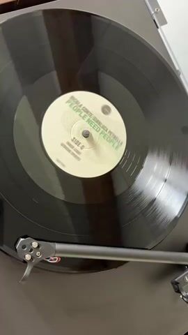 The video shows a black vinyl record spinning on a turntable. The record's label is white with green text that reads "NICOLA CONTE GIANLUCA PETRELLA PEOPLE NEED PEOPLE SIDE C UNDER LIGHT AFRIKA SPIRITS". The turntable has a dark gray base and a black tonearm with a silver cartridge. The camera angle is an overhead shot, providing a clear view of the record and turntable.