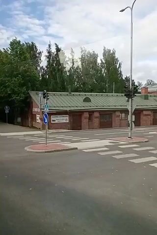 This video shows a view from a vehicle traveling through a street with a park-like setting. The scene is set in Kuopio, Finland. It appears to be daytime, with a cloudy sky.

The main subject is an "AUTOMUSEO" (Automobile Museum), a brick building with a green roof, visible on the right side of the road. Signs for the Automuseo are displayed on the building. Next to the museum, there are trees and a pedestrian crossing. Traffic lights and road signs, including a blue sign with a white pedestrian symbol and a downward-pointing blue arrow, are also visible.

On the left side of the road, there are more trees and a blue circular sign with a white symbol. The road is paved, and there are pedestrian crossings with white stripes. The presence of trees, grassy areas, and paved pathways suggests a well-maintained urban environment, possibly near a park or recreational area.

The video captures the movement of the vehicle, indicated by the blurring of the trees and the general flow of the scene. No people are clearly visible in the frame. The overall impression is that of a calm, ordinary day in a Finnish town.