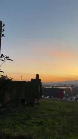 The video presents a landscape scene at dusk or dawn, featuring a mix of natural and urban elements. In the foreground, there's an old, overgrown ruin of a building, possibly made of stone. The building is covered in vegetation, obscuring much of its structure. The immediate ground is covered in green grass. Behind the ruins, the landscape transitions into a more industrial or commercial area with several large, modern buildings. These buildings appear to be warehouses or large retail structures, possibly including a store with a red bullseye logo. Beyond the buildings, in the distance, there's a line of hills or low mountains silhouetted against the sky. The sky itself is the focal point of the scene, showcasing a gradient of colors from a pale blue at the top to hues of orange and yellow near the horizon, indicating either a sunrise or sunset.