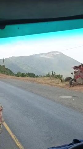 The video is taken from inside a vehicle, likely a truck, driving on a paved road. Through the windshield, a large, rounded mountain is visible in the distance. The mountain is mostly covered in green vegetation, but a lighter-colored area is visible near its peak. The road is wet and has a yellow line marking the edge. Some utility poles are visible along the side of the road. A pile of tires sits on the shoulder of the road. The vehicle is moving, and the scenery is passing by.