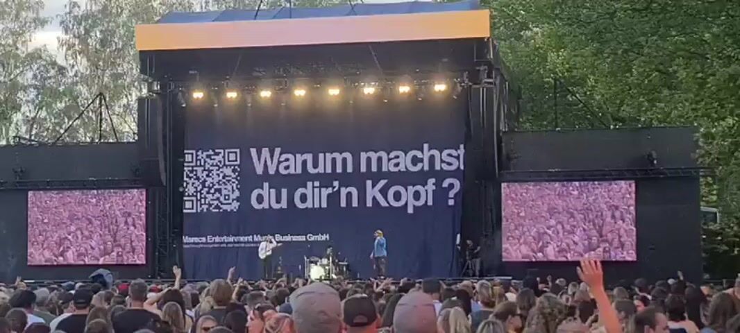 The video shows a large outdoor concert stage with a huge black banner in the background. The banner displays a QR code on the left and large white text on the right that reads "Warum machst du dir'n Kopf?". Below the text, in smaller print, it says "Moross Entertainment Music Business GmbH". Two musicians are visible on stage, one playing a guitar and the other standing near a drum set. Another person, likely a singer, is standing in front of the banner with a microphone. The stage is illuminated by bright lights, and two large screens on either side of the banner show blurred images of a crowd. The foreground is filled with a dense crowd of concertgoers, many with their hands raised. The overall environment appears to be a festival or outdoor concert event. A sign above the stage indicates "STARnacht am Neusiedler See 28. & 29. Mai 27".