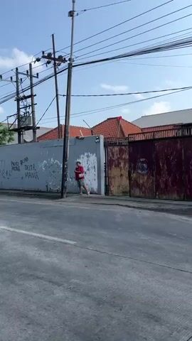 A person wearing a red shirt and white shorts stands near a gray wall with graffiti. The wall has a rusted metal gate to its right. Overhead, power lines crisscross a blue sky. The ground is asphalt.