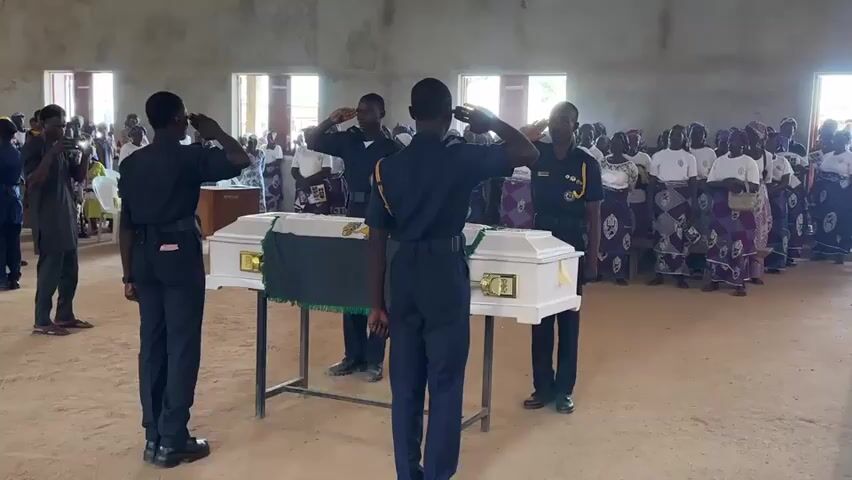 The video shows a solemn ceremony taking place inside a large room with a concrete floor and ceiling. The room appears to be a community hall or church with several windows. In the foreground, four individuals in dark uniforms with light-colored accents are standing at attention and saluting. They are positioned around a white coffin with gold handles, which rests on a metal table covered with a green cloth. A dark green blanket is draped over the coffin. Behind the coffin, a crowd of people, mostly women wearing traditional patterned fabric skirts and white tops, are gathered, also appearing to be attentive to the ceremony. Some people in the crowd are holding up their phones, presumably to record the event. To the left of the scene, another individual in a dark uniform is speaking into a microphone. The overall atmosphere is somber and respectful.