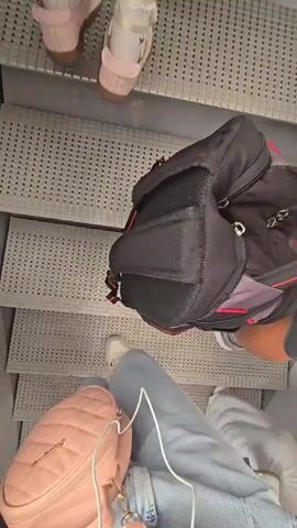 The video begins with a close-up shot of a child's feet in pink sandals ascending an escalator. The camera then pans up to show other passengers with luggage also on the escalator. One person is wearing a blue t-shirt and a gray Under Armour baseball cap. Outside the escalator, a pink airplane is visible on the tarmac. The video concludes with a shot of a person's hand resting on their thigh.