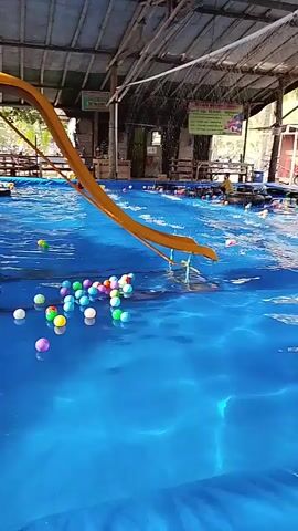 A wide-angle shot shows a shallow blue swimming pool with a yellow and orange slide on the left. The pool is filled with colorful plastic balls. A person wearing a green shirt is standing in the water near the slide. In the background, there are tables and chairs under a thatched roof structure, and more people are visible walking around and sitting. The environment appears to be an outdoor recreational area.