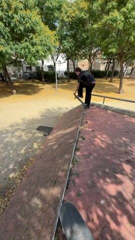 A young man with tattoos on his arm and wearing black pants and a black t-shirt performs a skateboarding trick. He rides his skateboard up a red brick ledge and then rides down a slope. He continues to ride on a dirt path and then stops his skateboard and picks it up. The scene is outdoors with trees and fallen yellow leaves on the ground. There is a metal railing in the background and parked cars.